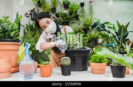 Asiatische Gärtner Frau trägt Gesichtsmaske und Schürze mit Schaufel, um Zimmerpflanzen und Kaktus zu transplantieren und sich um Zimmerpflanzen zu Hause, Hobby oder zu kümmern Stockfoto