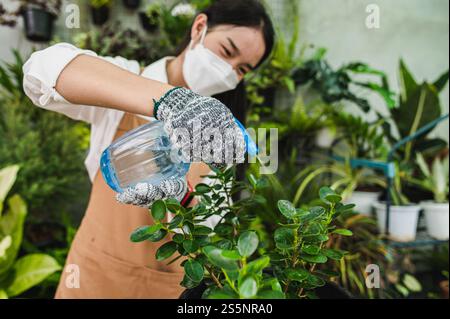 Selektiver Fokus, asiatische Gärtnerin Frau trägt Gesichtsmaske und Schürzenspray, um grüne Zimmerpflanze zu gießen, während kümmern Sie sich um Zimmerpflanze auf dem Tisch bei Stockfoto