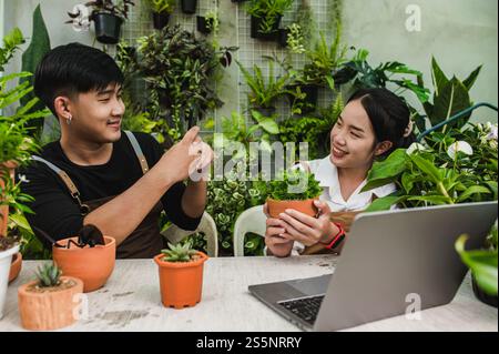 Junger Gärtner Mann mit Schürze benutzen Smartphone machen ein Foto seiner Freundin, sie hält Hauspflanze und lächelt mit glücklich zusammen Stockfoto