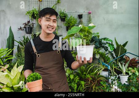 Portrait junger asiatischer Gärtner Mann stehend und hält zwei schöne Zimmerpflanze in Händen, er lächelt und sieht Pflanze mit glücklich aus Stockfoto