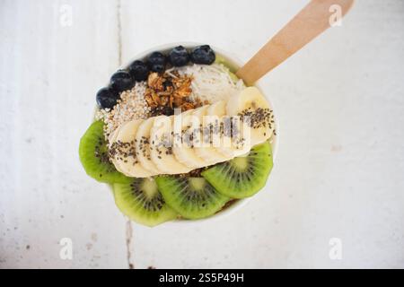 Eine Acai-Schüssel mit Blaubeeren, Chiasamen, Kiwis, Müsli und Kokosnuss befindet sich auf einem weißen Holztisch mit einem Holzlöffel Stockfoto