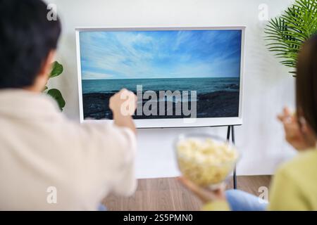 Asiatische Familie, die gemeinsam Smart TV ansieht und mit der Fernbedienung Popcorn zu Hause hält, während sie mit der Fernbedienung Filme oder TV-Serien ausgibt Stockfoto