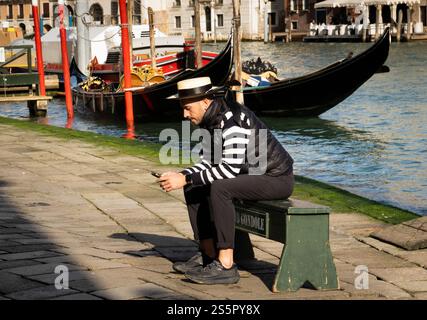 Ein Gondoliere sitzt mit seinem Handy an einem Kanal in Venedig, Italien. Stockfoto