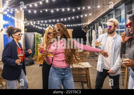 Glückliche moderne afrikanische Frau mit langen geflochtenen Haaren, die mit multiethnischen Freunden bei einer Nachtparty draußen auf der Terrasse einer Bar tanzen Stockfoto