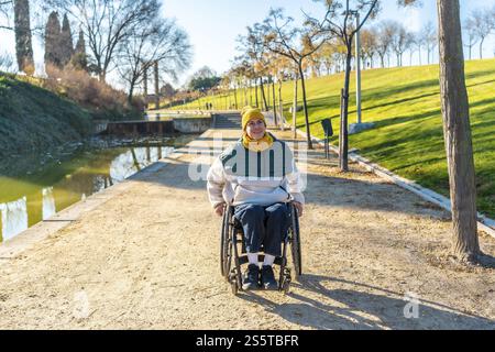 Glückliche junge Frau mit Rollstuhl, die einen sonnigen Tag im Park in der Nähe eines kleinen Sees genießt, was die barrierefreie Erholung und Inklusivität fördert Stockfoto