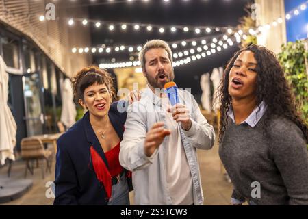 Ein Mann und zwei Frauen mit verschiedenen Rassen singen Karaoke und haben Spaß bei einer Abendparty auf der Terrasse einer Bar Stockfoto