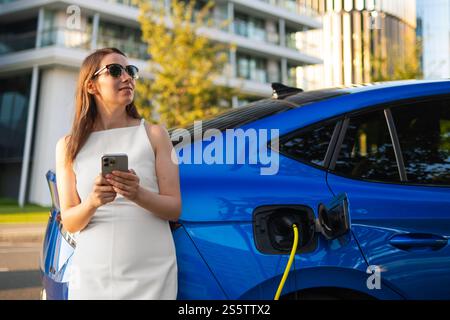 Lächelnde Frau in einem weißen Kleid benutzt ein Smartphone, während sie draußen ein Elektrofahrzeug auflädt Stockfoto