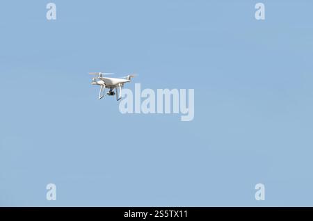 Große weiße Drohne, die in einem hellen wolkenlosen blauen Himmel schwebt, Funksteuerhubschrauber mit tragbarer Videokamera-Versorgung. Große weiße Drohne, die in A schwebt Stockfoto