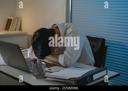 Ein müder asiatischer Geschäftsmann, der allein am Tisch einschlief und nach Nachtarbeit den Kopf an den Händen hielt. asiatischer männlicher Mitarbeiter. Stockfoto