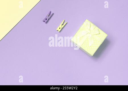 Kleine gelbe Geschenkbox und zwei Stifte liegen auf Textur Hintergrund von Mode pastellgelb und violett Farben Papier in minimalem Konzept. Kleines gelbes Geschenk Stockfoto