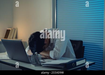Ein müder asiatischer Geschäftsmann, der allein am Tisch einschlief und nach Nachtarbeit den Kopf an den Händen hielt. asiatischer männlicher Mitarbeiter. Stockfoto
