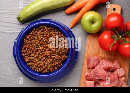 Schüssel mit trockenem Haustierfutter, rohem Fleisch und Naturprodukten auf farbigem Holzhintergrund Stockfoto