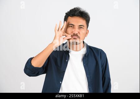 Porträt eines jungen asiatischen Mannes in blauem Hemd, der ein Geheimnis auf weißem Hintergrund isoliert hält. Ausdruck und Lifestyle-Konzept. Stockfoto