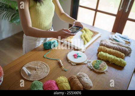 Asiatische Frau macht Smartphone Foto Punch Needle. Telefonpost in sozialen Netzwerken in der Studiowerkstatt. Designer-Arbeitsplatz Handgefertigtes Bastelprojekt DIY Stockfoto