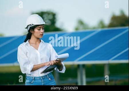 Porträt Asiatische junge Ingenieurin, die einen Schutzhelm trägt, der vor dem Sonnenkollektor steht, um den Betrieb in Papieren zu überprüfen, während sie arbeiten Stockfoto