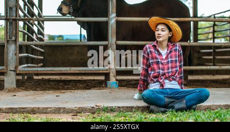 Porträt einer müden jungen asiatischen Bäuerin, die auf dem Boden im Kuhstall sitzt. Landwirtschaft, Landwirtschaft, Menschen, Technologie und Tierhaltung Stockfoto
