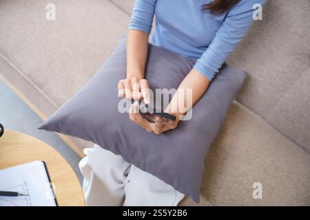 Asiatische Frau, die Smart Home kontrolliert. Mobiltelefon mit Smart-Home-App im Wohnzimmer. Touchscreen-Ai-Technologie. Fernbedienungssystem für Zuhause Stockfoto