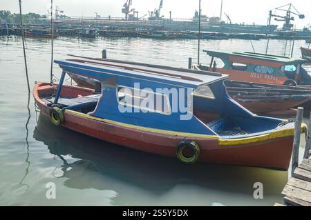 Ein kleines blaues hölzernes Fischerboot, das sich auf einem hölzernen Pier mit einem Industriegebiet und ruhigem Meer im Hintergrund lehnt. Seitenansicht, Indonesien, 14. Juni 2023 Stockfoto