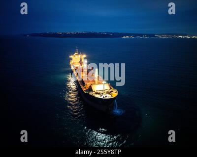 Beleuchtetes Frachtschiff auf See bei Nacht, von oben gesehen mit Küste und leuchtenden Lichtern der Stadt in der Ferne Stockfoto