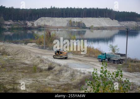 Caterpillar-Lader und Muldenkipper arbeiten im Tagebau. Schwere Maschinen in der offenen Grube am Fluss. Planierschild und Lkw beim Graben und Stockfoto