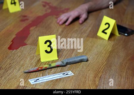 Tatortuntersuchung - blutiges Messer und Opferhand mit gelben kriminellen Markierungen auf Küchenboden. Mordbeweise. Tatortuntersuchung Stockfoto