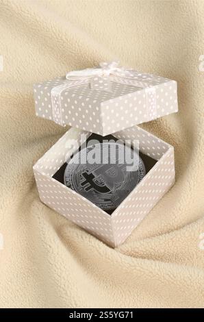 Der silberne Bitcoin liegt in einer kleinen orangen Geschenkbox mit einer kleinen Schleife auf einer Decke aus weichem und flauschigem hellorangen Fleecestoff mit einem großen Stockfoto