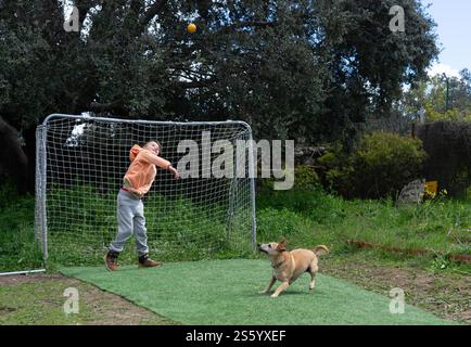 Kind, das mit Hund und Ball im Hinterhof spielt: Ball auf Kleinhund durch Fußballtor werfen Stockfoto