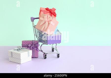 Kleine Geschenkboxen in verschiedenen Farben mit Bändern liegen im Warenkorb auf einem violetten und blauen pastellfarbenen Hintergrund. Kleine Geschenkboxen in verschiedenen Farben Stockfoto