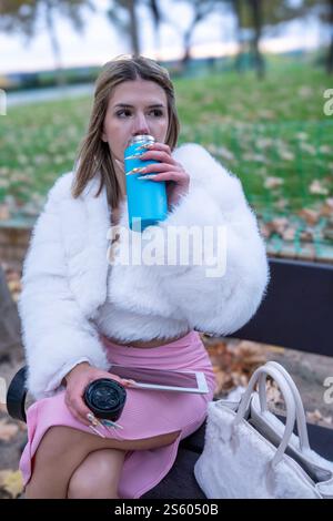 Junger Influencer trinkt aus wiederverwendbarer Wasserflasche, während er auf einer Bank im Park sitzt und Tablette und Kaffeetasse hält Stockfoto