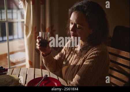 Ein ruhiger Morgenmoment. Eine Frau, die ein Glas Saft hält, während sie weiches Sonnenlicht genießt, das durch das Fenster in der Nähe eines Tellers mit Crepes hereinkommt. Stockfoto