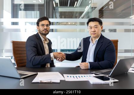 Zwei ernste Kollegen, die die Hand schütteln. Ernsthafte und selbstbewusste Männer in Businessanzügen sitzen an einem Konferenztisch und schauen in die Kamera Stockfoto