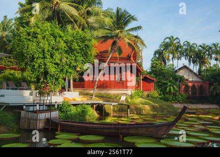 Ein Holzhaus im thailändischen Stil mit einem roten Dach, umgeben von tropischer Vegetation, einem Teich mit Lilienpads und einem hölzernen Langboot im warmen Sonnenlicht. Stockfoto