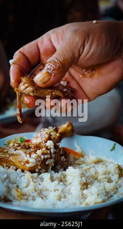 Eine Hand, die ein Stück Huhn über einem Teller Reis hält und das traditionelle Essen mit den Händen zeigt. Stockfoto