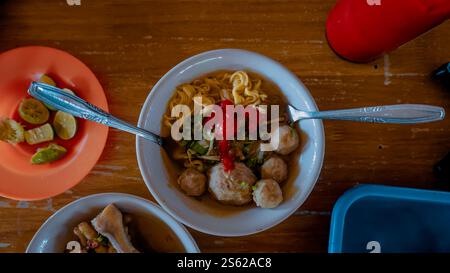 Eine Schüssel Nudelsuppe mit Fleischbällchen, garniert mit roter Sauce und Kräutern, auf einem Holztisch. Ein Teller mit Kalkkkeilen und eine andere Schale sind teilweise V Stockfoto