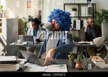 Junger Arbeiter in Clown-Perücke, der die Escape-Taste auf der Tastatur des Laptops drückt, während er Kaffee trinkt und auf den Bildschirm blickt Stockfoto