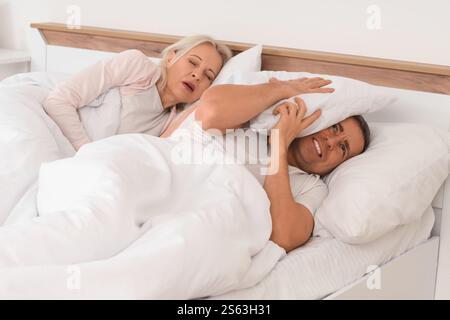 Gereizter reifer Mann, der mit seiner schnarchenden Frau im Schlafzimmer liegt Stockfoto