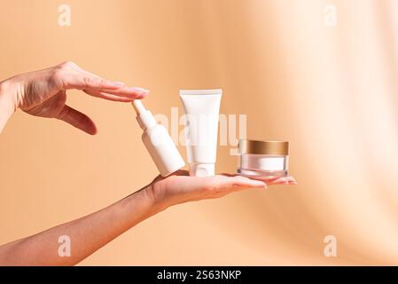 Frauenhände halten weiße Kosmetikbehälter, Gesichtscremebecher, Maskenröhrchen, Serumflasche, Lotion auf pastellorangen Hintergrund, Kopierraum. Hautpflege Co Stockfoto