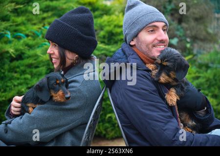 Junges Paar, das Mützen trägt, sich im Freien auf Stühlen zurücklehnt, ihre kleinen Hunde hält, lächelt und wegblickt Stockfoto