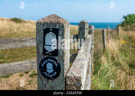 Eine Markierung entlang des England Coastal Path steht an einem hölzernen Pfosten, umgeben von hohem Gras und dem ruhigen Meerblick Stockfoto