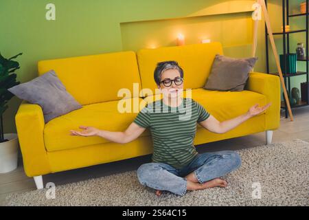 Ganzkörperfoto einer netten pensionierten Frau Sitzen Boden Meditation tragen grüne Kleidung Genießen Sie die gemütliche Innenwohnung drinnen Stockfoto