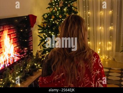 Die junge Frau, die in eine festliche Decke gewickelt ist, genießt die gemütliche Weihnachtsatmosphäre in einem warm beleuchteten Wohnzimmer Stockfoto