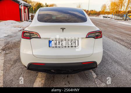 Rückansicht des weißen Tesla Model Y, der im Winter unter Abendsonne auf einer verschneiten Straße in der Nähe des roten Gebäudes geparkt ist. Schweden. Stockfoto