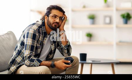 Gelangweilter indischer Kerl, der mit Joystick auf der Couch sitzt Stockfoto