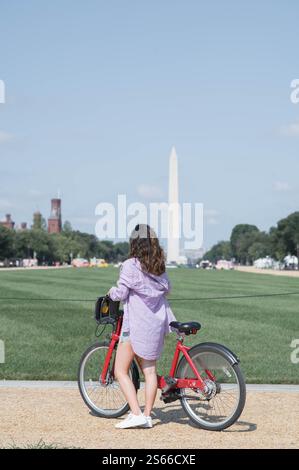 An einem sonnigen Sommertag im Nationalpark in Washington DC hat ein Mädchen aufgehört, ihr rotes Fahrrad zu fahren, um sich das Washington Monument Memorial anzusehen. Stockfoto