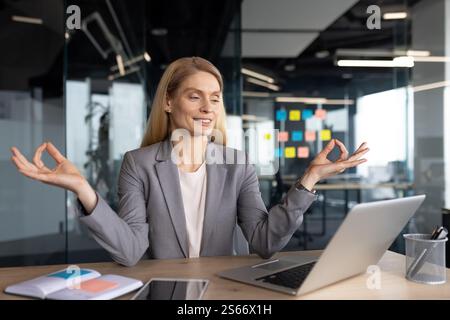 Fröhliche Frau, die in Lotusposien im Büro am Arbeitsplatz meditiert. Reife Geschäftsfrau im Business-Anzug, erholt und entspannt sich beim Online-Video-Yoga-Kurs mit Laptop. Stockfoto