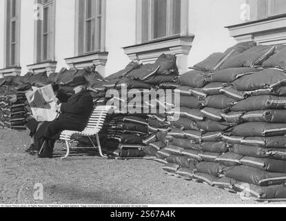 In den 1940er Jahren Ein älteres Ehepaar sitzt auf einer Bank und liest eine Zeitung vor einem Gebäude mit Sandsäcken, die sich vor den Kellerfenstern stapeln. Dies wurde durchgeführt, um das Gebäude und alle Personen im Inneren zu schützen, falls ein Bombenangriff stattfinden würde. Das Bild wurde am 12. april 1940 während des Zweiten Weltkriegs aufgenommen. Stockfoto