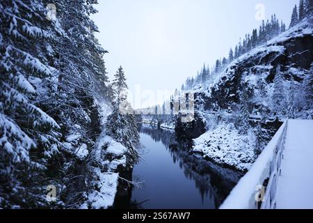 Eine friedliche Winterszene mit einer verschneiten Waldschlucht mit einem ruhigen, reflektierenden Fluss. Ein schneebedecktes Brückengeländer führt das Auge durch die ruhige Landschaft, die satinierten Bäume und die zerklüfteten Klippen. Stockfoto