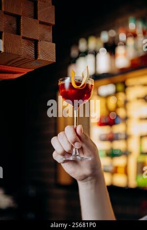 Hand hält einen roten Cocktail mit orangefarbenen Verzierungen vor einem lebhaften Barhintergrund Stockfoto