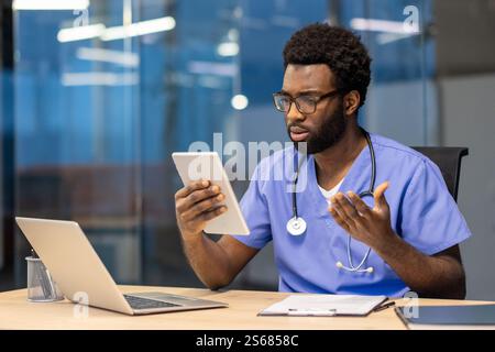 Junger erfahrener Arzt mit Tablet-Computer, der am Schreibtisch sitzt, mit Kollegen spricht und Patienten aus der Ferne konsultiert. Mann in blauem medizinischem Mantel und Stethoskop, der in der Klinik sitzt. Stockfoto