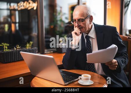 Konzentrierter Geschäftsmann in elegantem Anzug mit Brille mit Laptop und Untersuchung von Dokumenten, während er Kaffee trinkt und im Café sitzt. Stockfoto